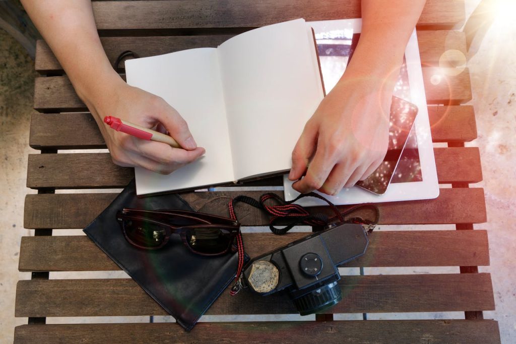 Top view of hands writing in notebook on wooden table with vintage camera and tablet.